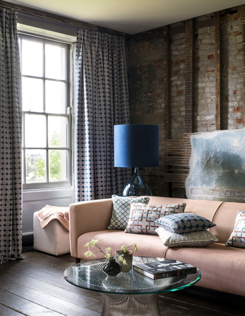 A peach fuzz sofa with dark blue lamp shade and patterned blue curtains. 