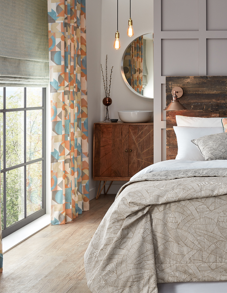 A comfy bedroom with grey bedding, patterned curtains and grey Roman blinds.