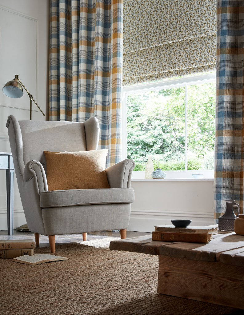 A comfortable sitting room with check curtains and a patterned Roman blind. There is a warm orange cushion on the cosy armchair.